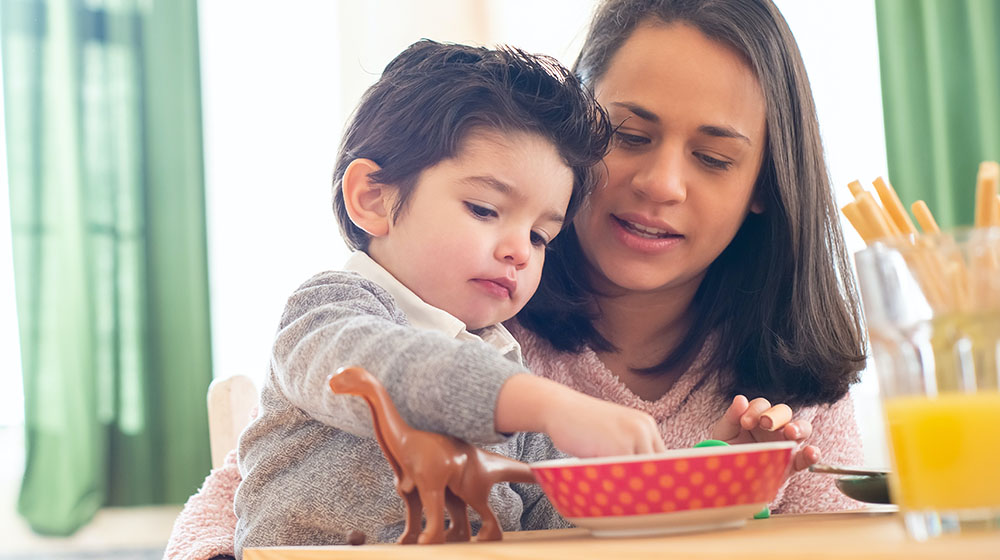 Boy feeding his mom a bite of cereal at the table with other siblings